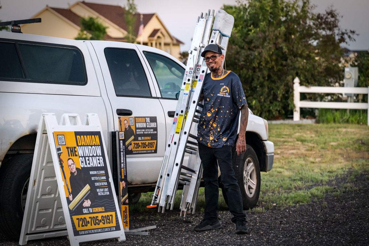 Damian The Window Cleaner standing beside branded truck and matching A-frame signs during residential job, showcasing consistent yellow and black branding, printed by WCR Marketing.