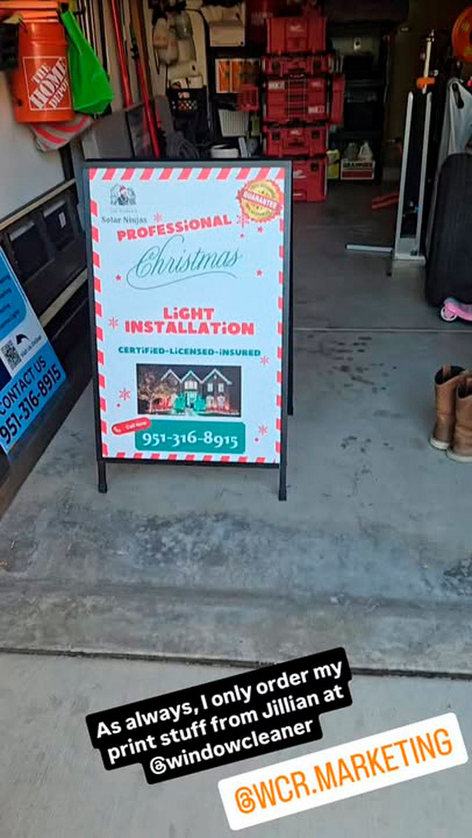 Christmas light installation A-frame sign inside garage promoting professional holiday lighting services with red and white candy-cane border, printed by WCR Marketing.