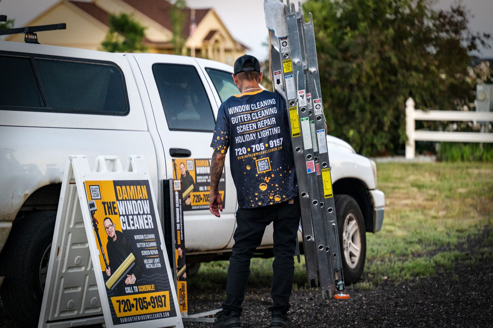Damian The Window Cleaner technician carrying a ladder beside a branded truck and A-frame signs in a neighborhood, printed by WCR Marketing.