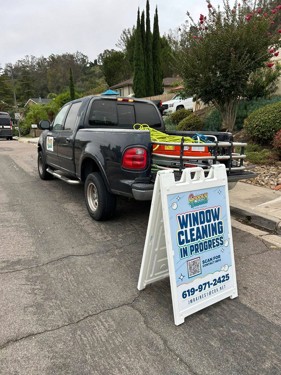 Groovy Bubbles Window Cleaning A-frame sign placed near parked truck and equipment on suburban street, promoting window cleaning in progress, printed by WCR Marketing.