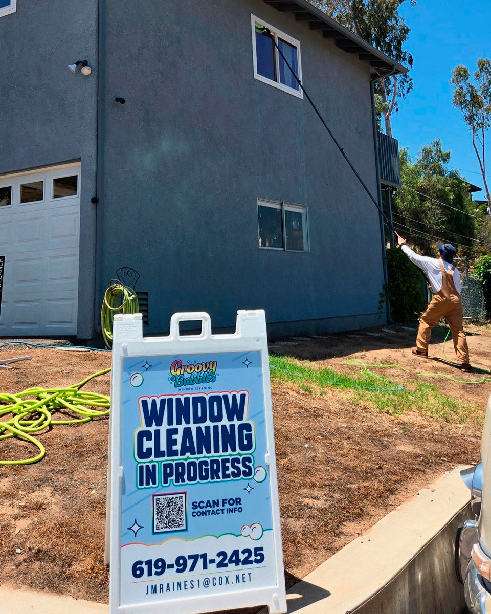Groovy Bubbles Window Cleaning A-frame sign at active jobsite with technician cleaning upper-story windows using extension pole, printed by WCR Marketing.
