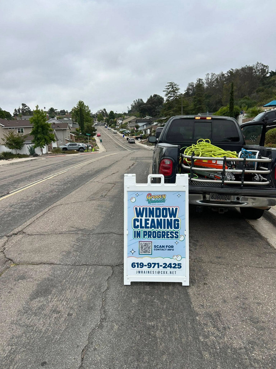 Groovy Bubbles Window Cleaning A-frame sign placed behind service truck on residential street with bright blue design, printed by WCR Marketing.
