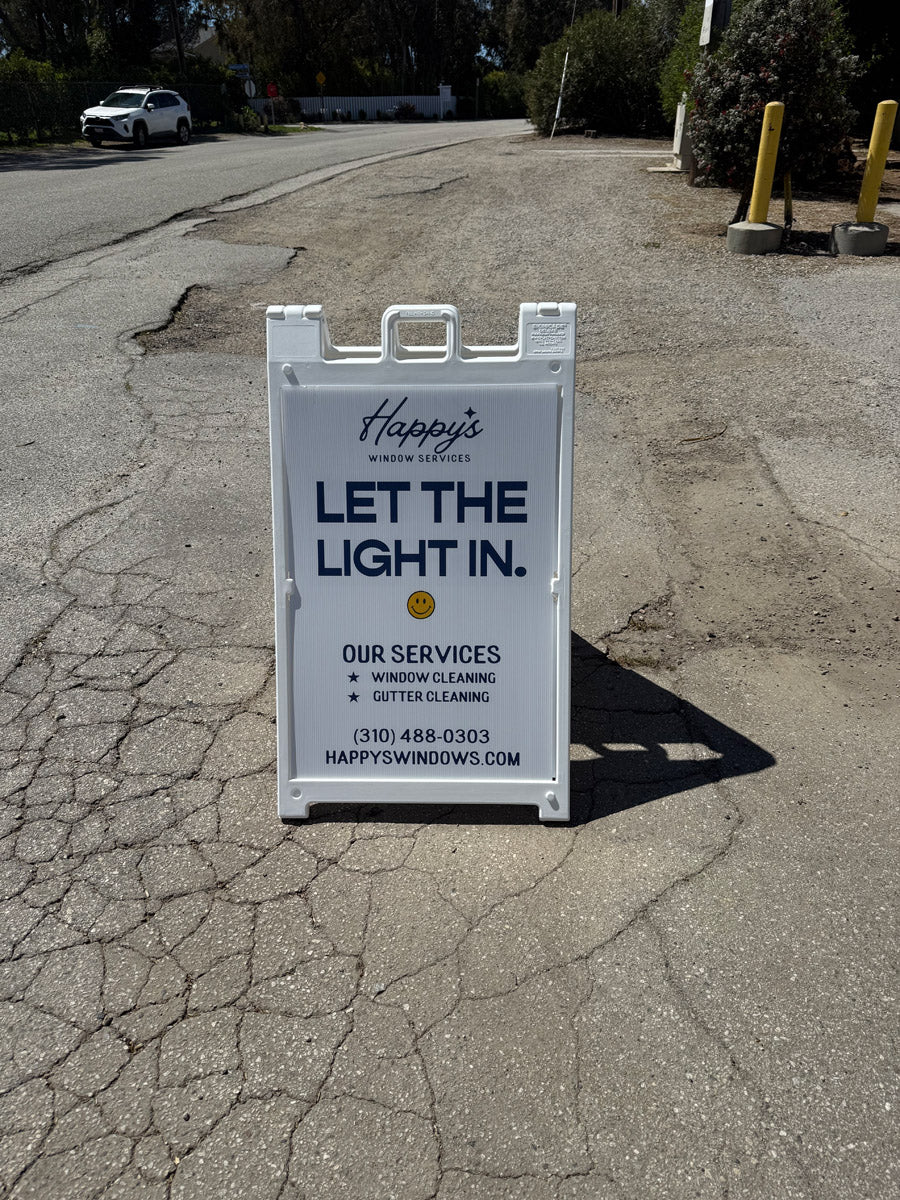 Happy’s Window Services A-frame sign on roadside reading “Let the Light In” promoting window and gutter cleaning services, printed by WCR Marketing.