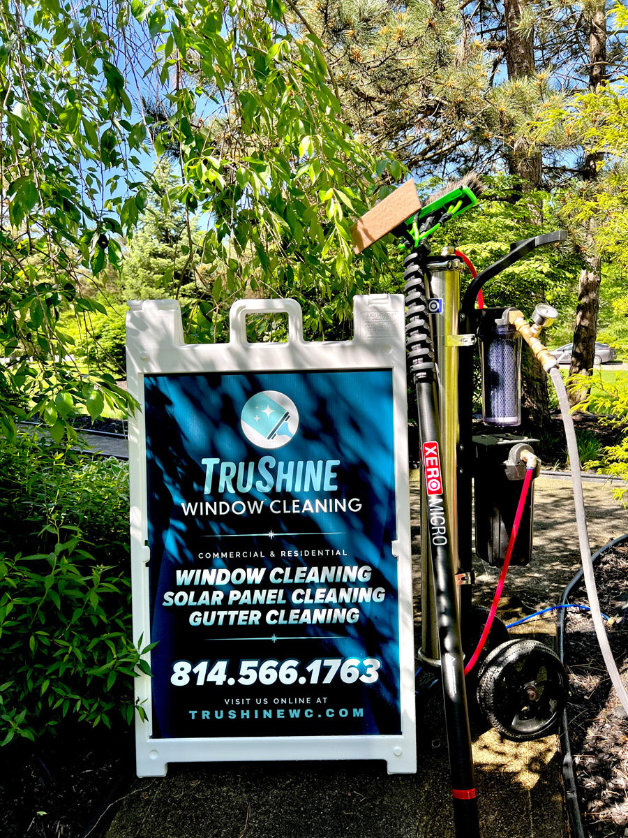 TruShine Window Cleaning A-frame sign displayed outdoors with XERO pole and purification system beside it, promoting solar panel and gutter cleaning, printed by WCR Marketing.