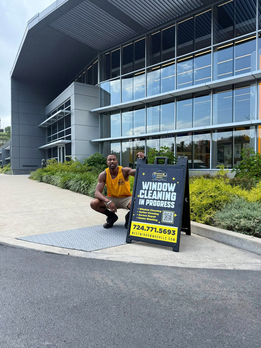 West Window Wash A-frame sign outside modern glass commercial building with cleaner posing beside it, printed by WCR Marketing.