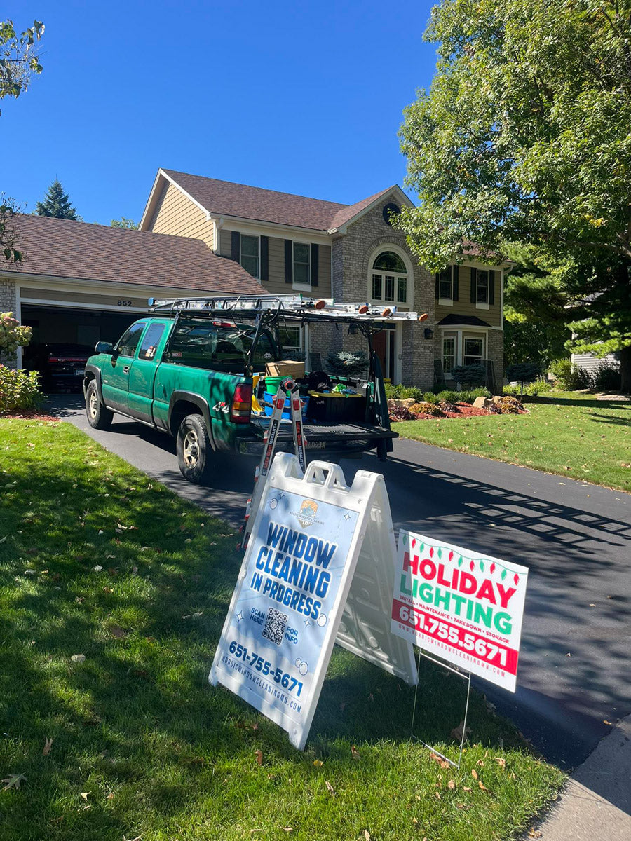 Green service truck parked in front of home with window cleaning A-frame sign and holiday lighting yard sign displayed on the lawn, printed by WCR Marketing.