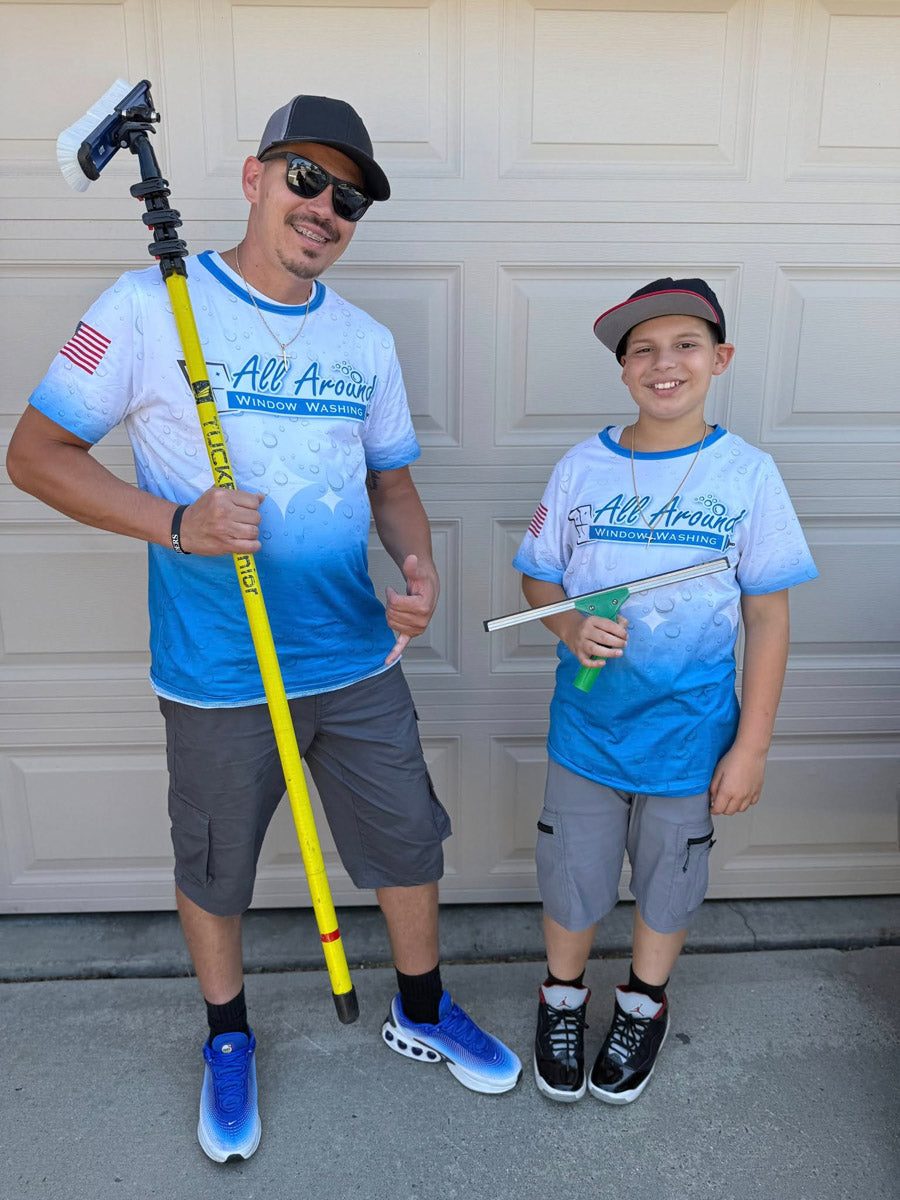 Father and son wearing matching All Around Window Washing dye-sub jerseys, holding a water-fed pole and squeegee in front of a garage, printed by WCR Marketing.