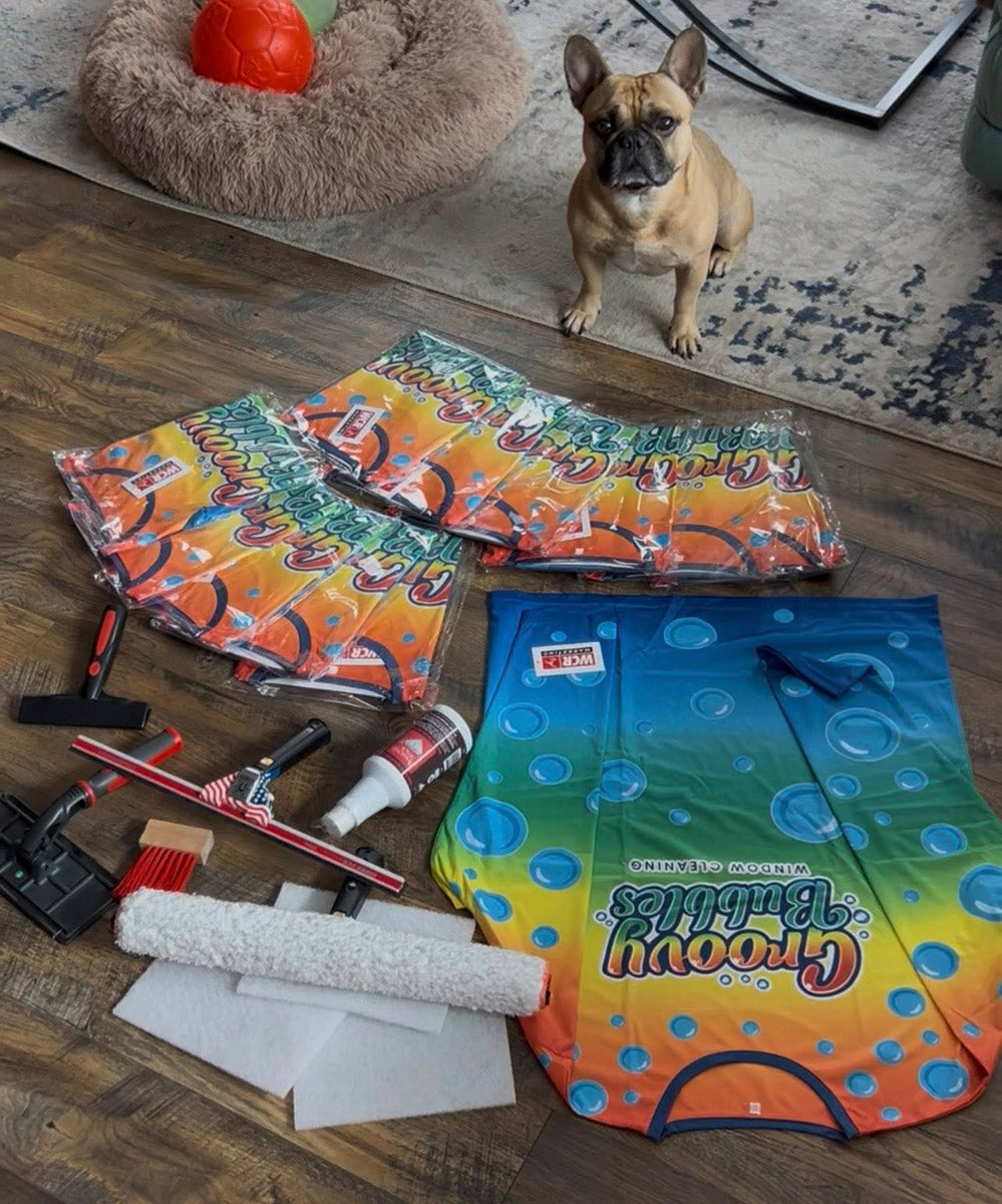 Groovy Bubbles Window Cleaning branded shirts with squeegees and cleaning tools arranged on wood floor beside small dog, printed by WCR Marketing.