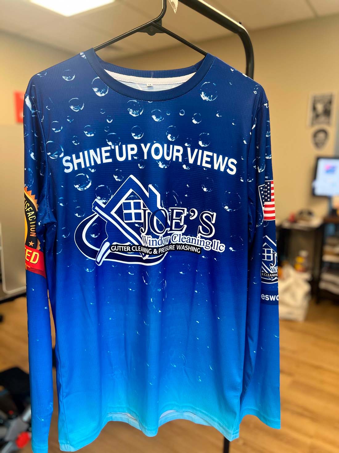 Joe’s Window Cleaning long sleeve dye-sublimated shirt reading “Shine Up Your Views,” featuring American flag sleeve patch, printed by WCR Marketing.