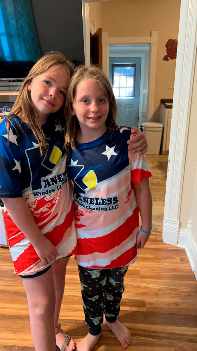 Two kids in matching Kaneless Window Cleaning patriotic dye-sublimated shirts, standing indoors and smiling, printed by WCR Marketing.