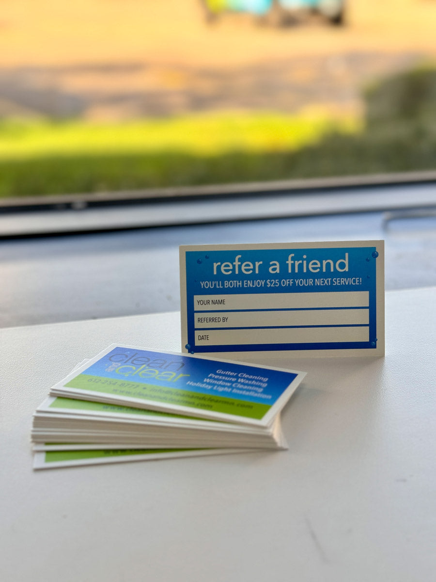 Clean and Clear referral card offering $25 off next service, displayed beside stack of business cards promoting window and gutter cleaning, printed by WCR Marketing.