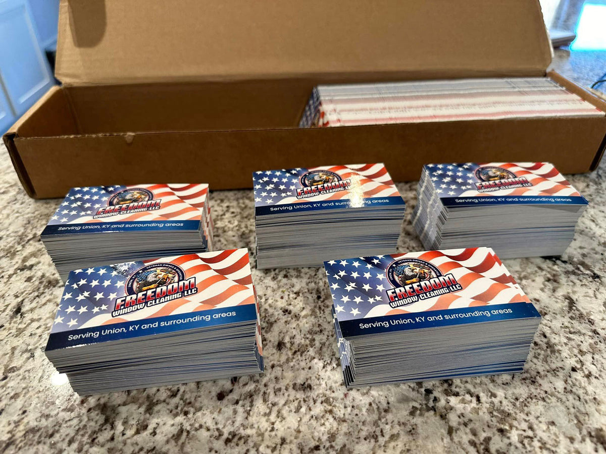 Freedom Window Cleaning business cards stacked in multiple piles on countertop, showcasing patriotic red, white, and blue theme, printed by WCR Marketing.