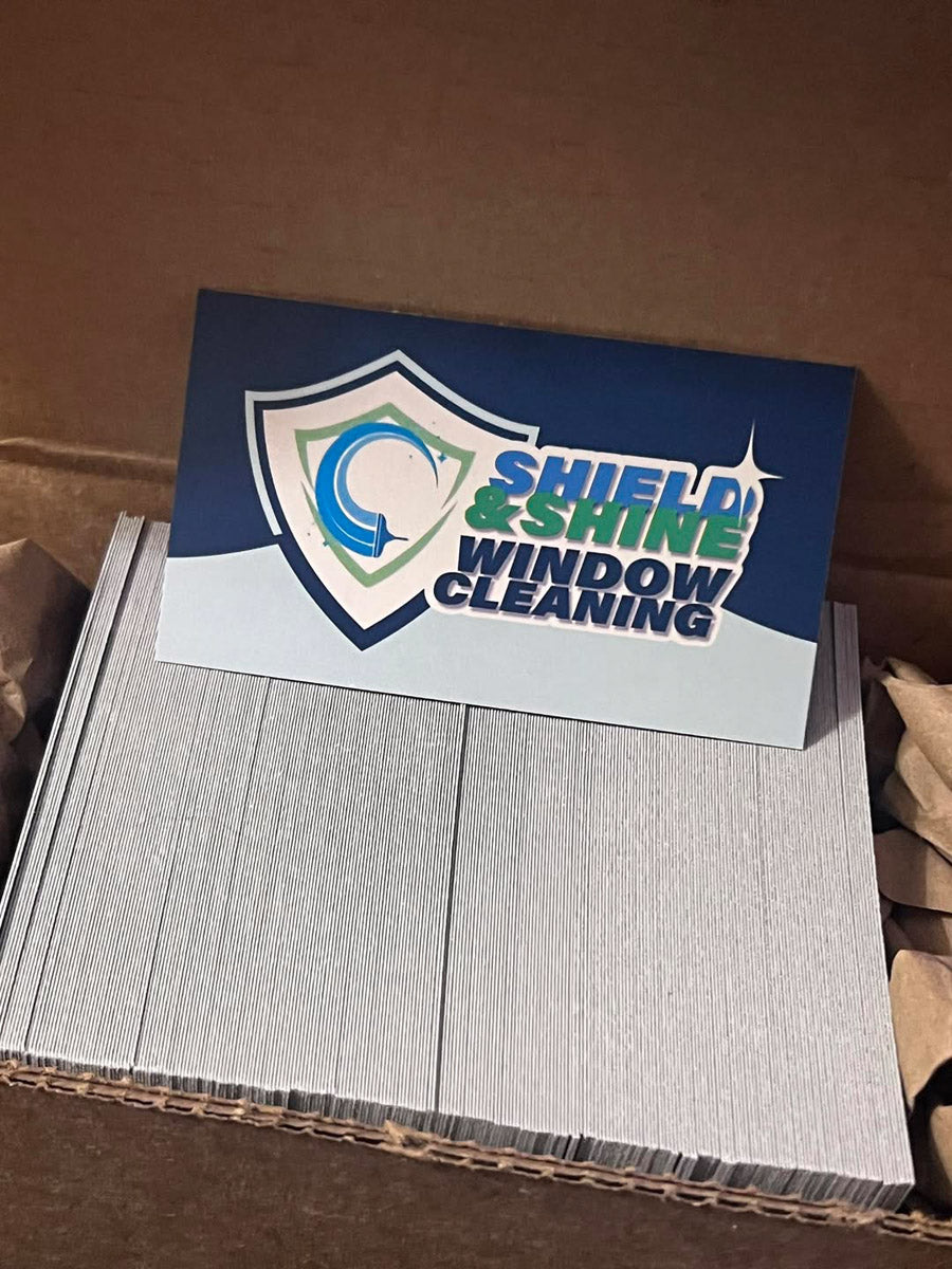 Shield and Shine Window Cleaning business cards featuring blue and green shield logo neatly stacked in shipping box, printed by WCR Marketing.