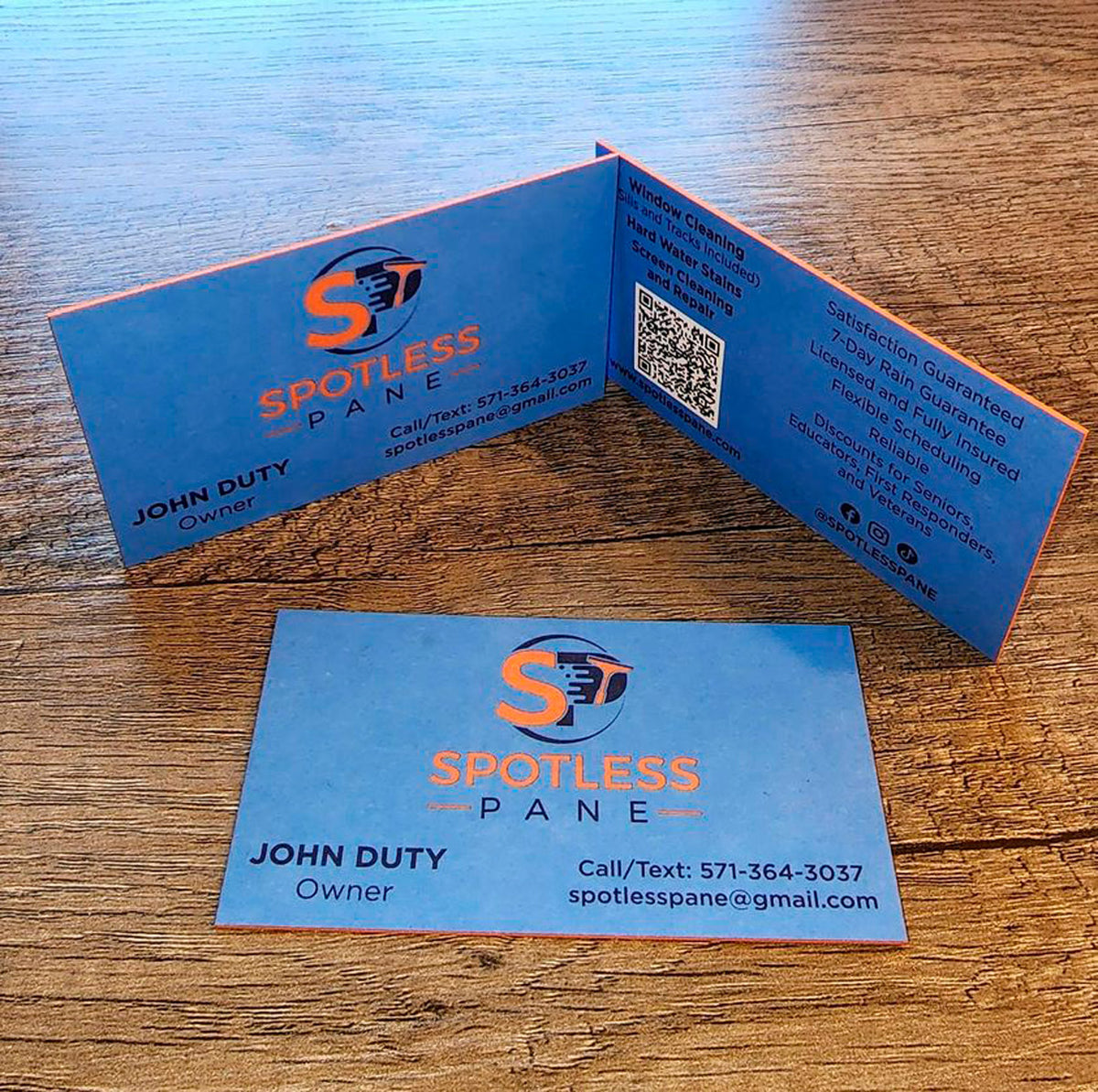 Spotless Pane  business cards on wooden surface featuring orange and blue branding and QR code for easy contact, printed by WCR Marketing.