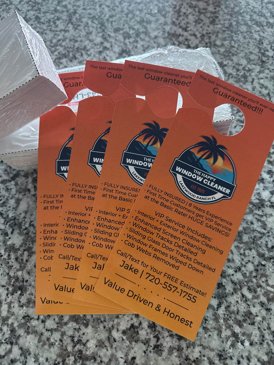 The Happy Window Cleaner orange VIP service door hangers stacked on countertop, featuring palm tree and sunset logo, printed by WCR Marketing.