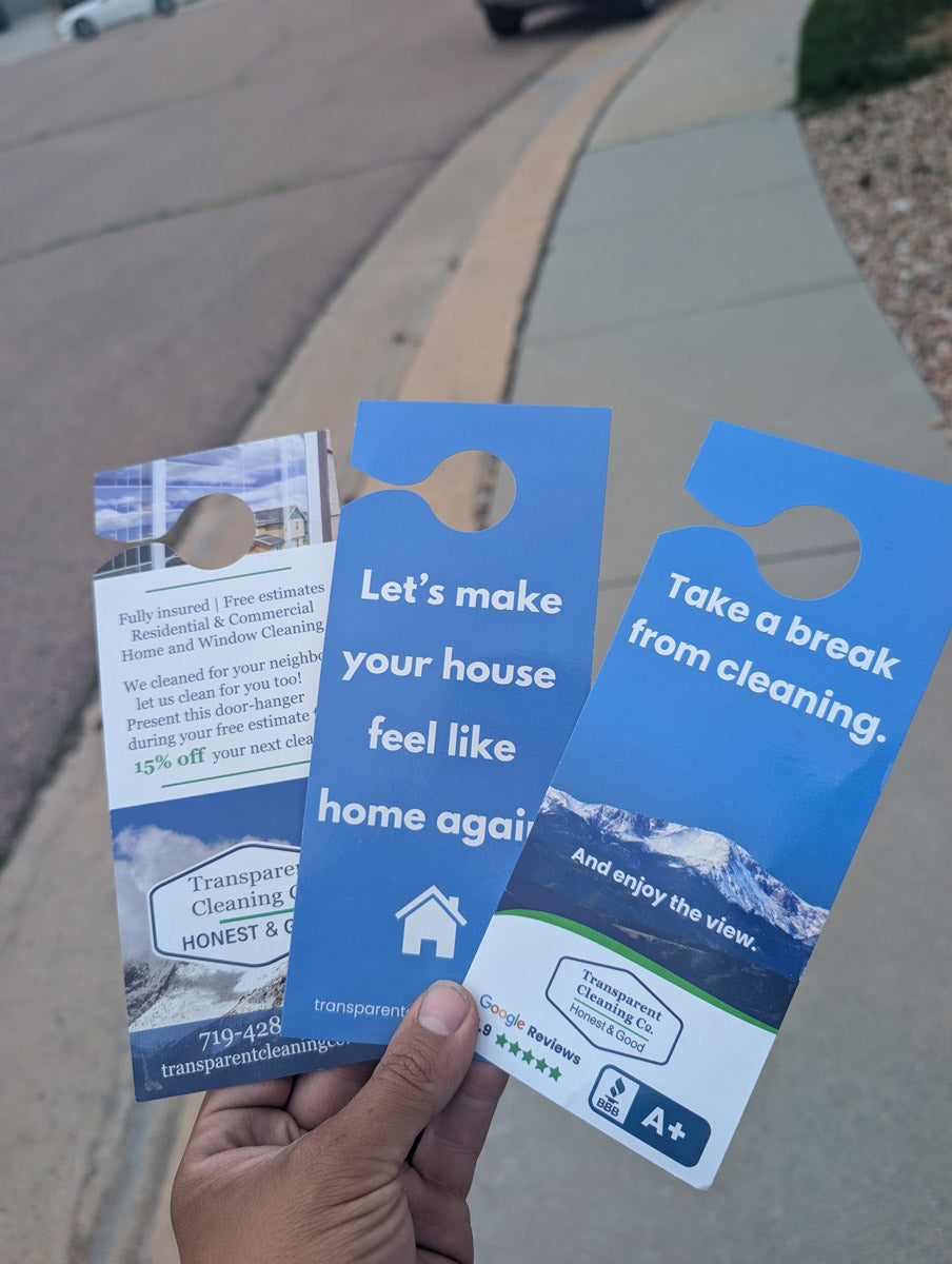 Transparent Cleaning Company door hangers in hand showing three versions with mountain imagery and blue motivational text, printed by WCR Marketing.