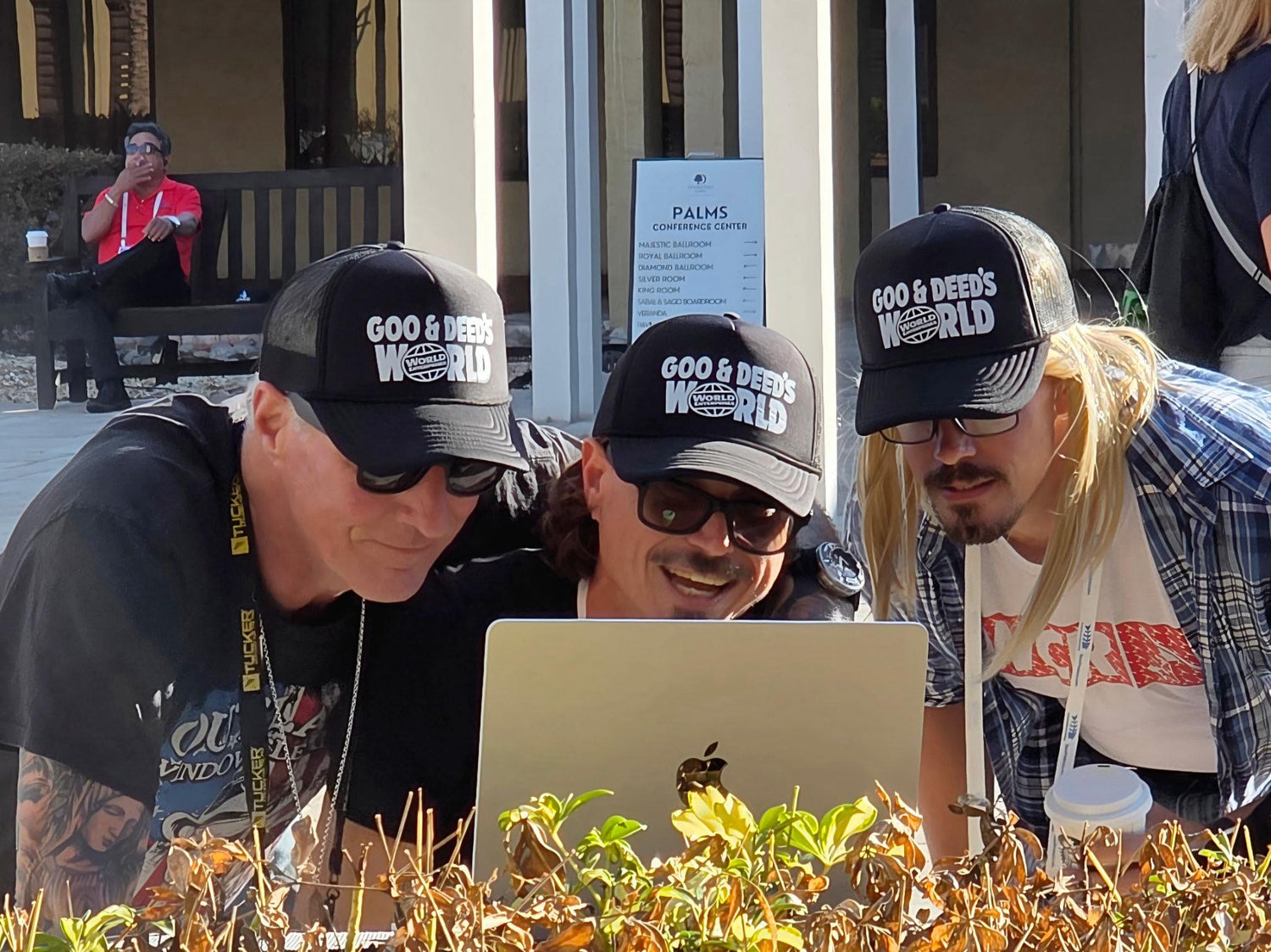 Three people wearing Goo and Deeds World Enterprises black trucker hats while looking at a laptop outdoors
