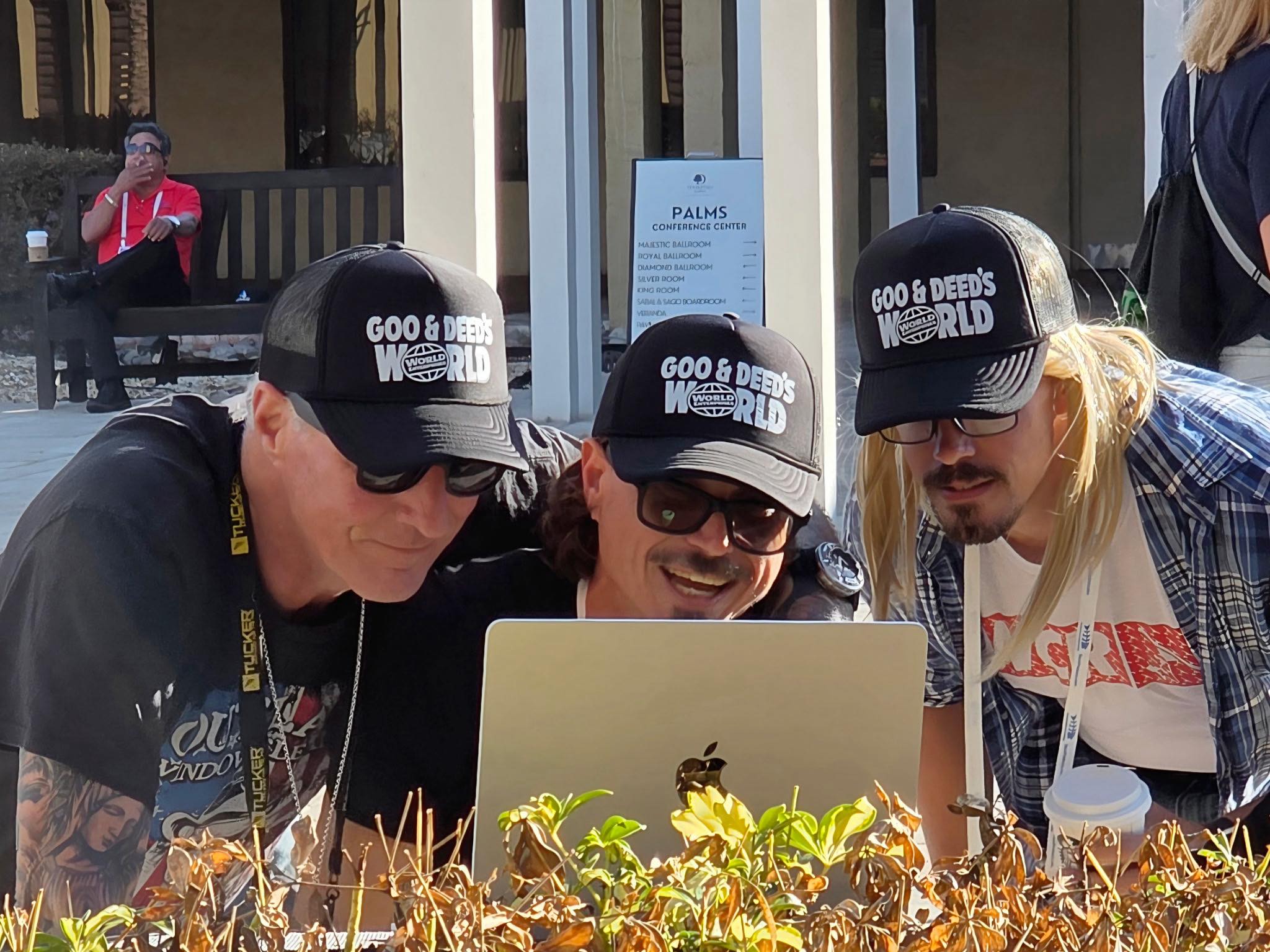 Three people wearing Goo and Deeds World Enterprises black trucker hats while looking at a laptop outdoors