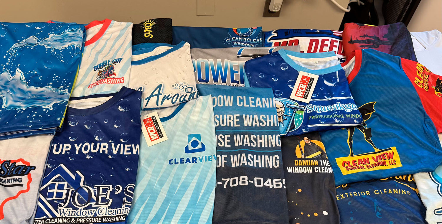 Assorted custom jerseys for multiple window cleaning and soft washing companies spread across a tabletop, printed by WCR Marketing.
