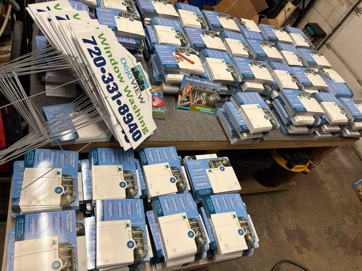 Stacks of Deja View Window Washing direct mail postcards and bundled yard signs spread across a shop table, printed by WCR Marketing.
