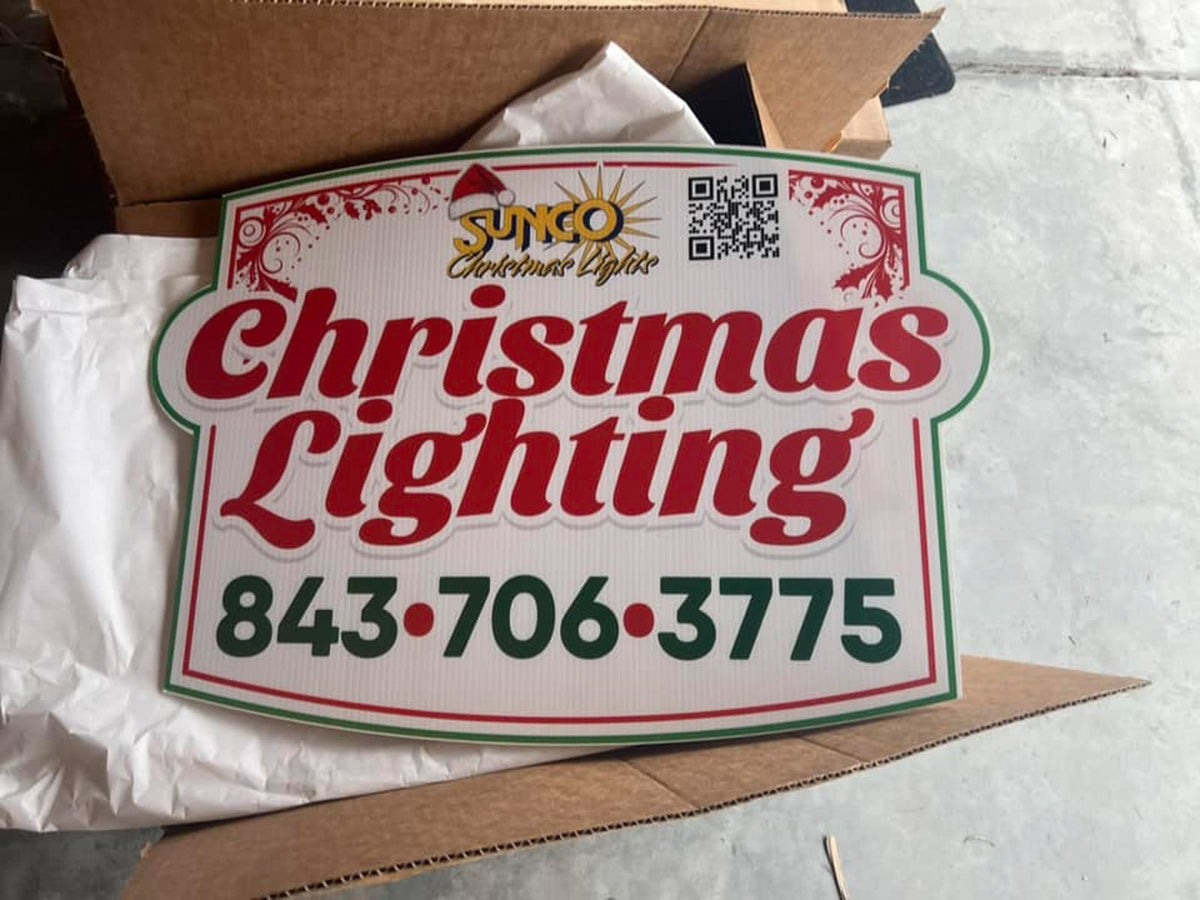 Sunco Christmas Lights die-cut yard sign with festive red and green Christmas lettering propped in an open box, printed by WCR Marketing.